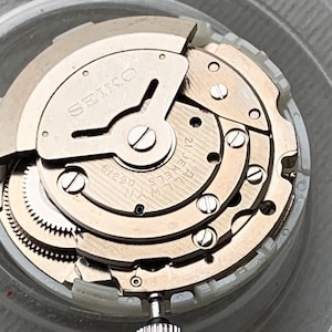 May include: Close-up of a watch movement with the brand name "SEIKO" visible. The mechanism features multiple gears, screws, and a crown. The watch movement is silver and gold in color, with the text "21 JEWELS" and "LW-N A6319" also visible.