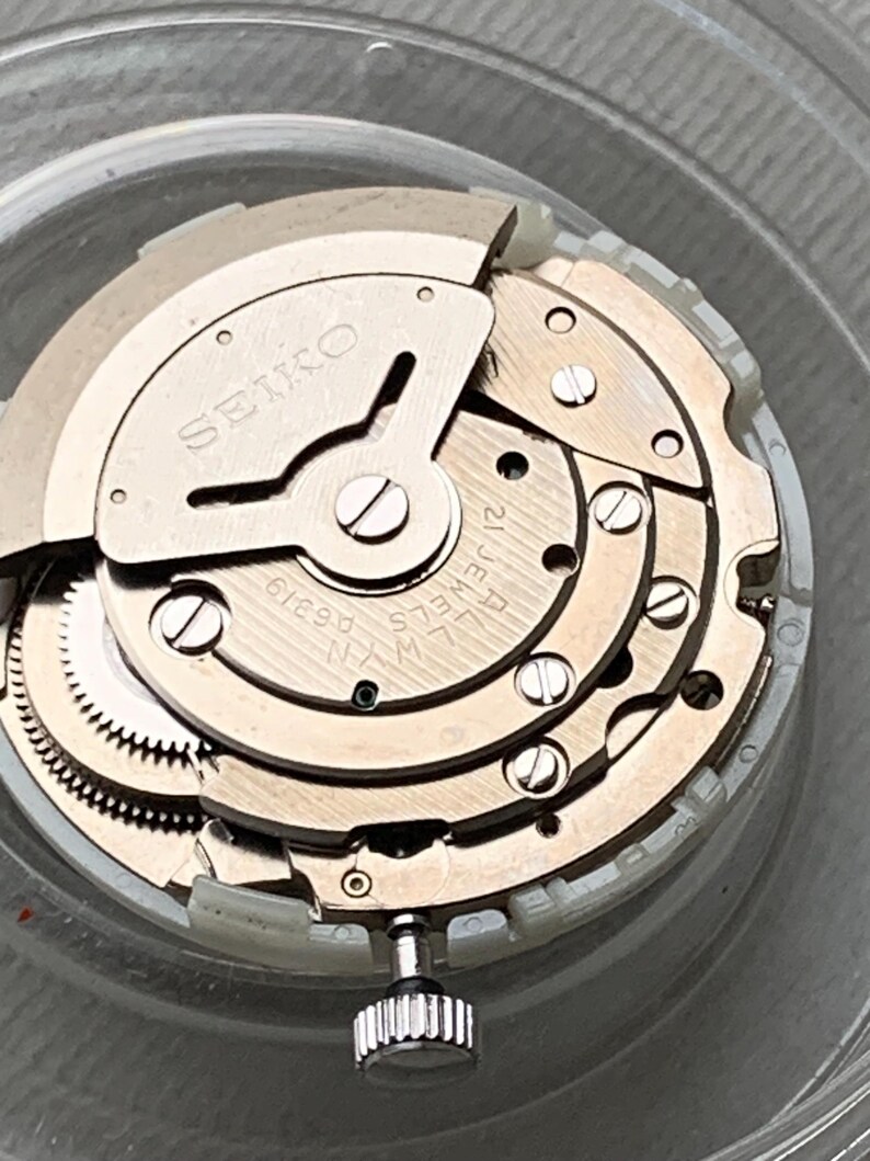 May include: Close-up of a watch movement with the brand name "SEIKO" visible. The mechanism features multiple gears, screws, and a crown. The watch movement is silver and gold in color, with the text "21 JEWELS" and "LW-N A6319" also visible.