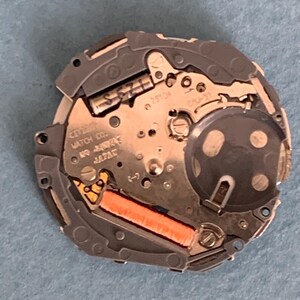 May include: A close-up of a Citizen watch movement. The watch mechanism is silver and gold with an orange coil. The text "CITIZEN WATCH CO." and "JAPAN" are visible. The watch movement is on a blue surface.