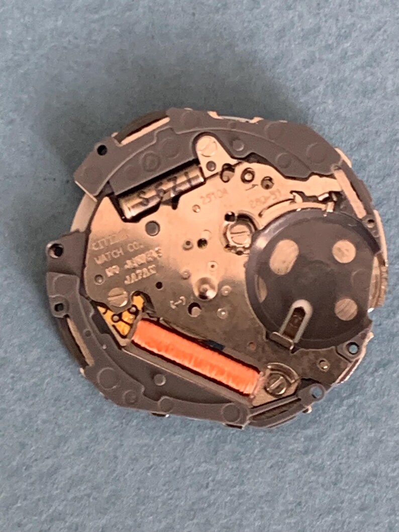 May include: A close-up of a Citizen watch movement. The watch mechanism is silver and gold with an orange coil. The text "CITIZEN WATCH CO." and "JAPAN" are visible. The watch movement is on a blue surface.