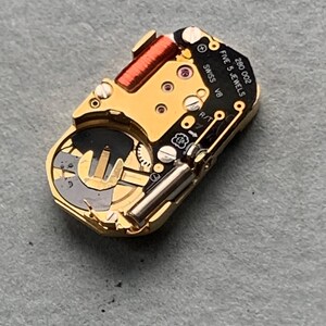 May include: Close-up of a gold-toned watch movement with intricate gears, a red coil, and the text "SWISS V8 FIVE 5 JEWELS 280 002". The watch mechanism is set against a gray surface, showcasing its detailed components.