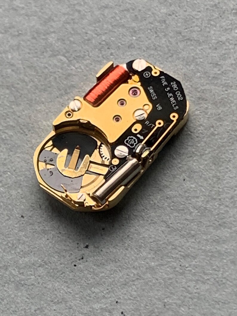 May include: Close-up of a gold-toned watch movement with intricate gears, a red coil, and the text "SWISS V8 FIVE 5 JEWELS 280 002". The watch mechanism is set against a gray surface, showcasing its detailed components.