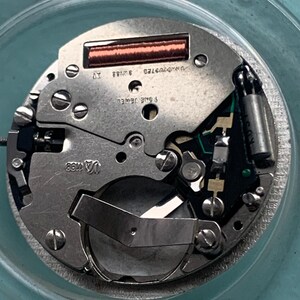 Op de afbeelding: Close-up van een uurwerk met zilverkleurige metalen componenten. Het uurwerk heeft een koperkleurige spoel, een groene printplaat en de tekst "SWISS" en "1 ONE JEWEL". Het uurwerk zit in een doorzichtige, ronde container.