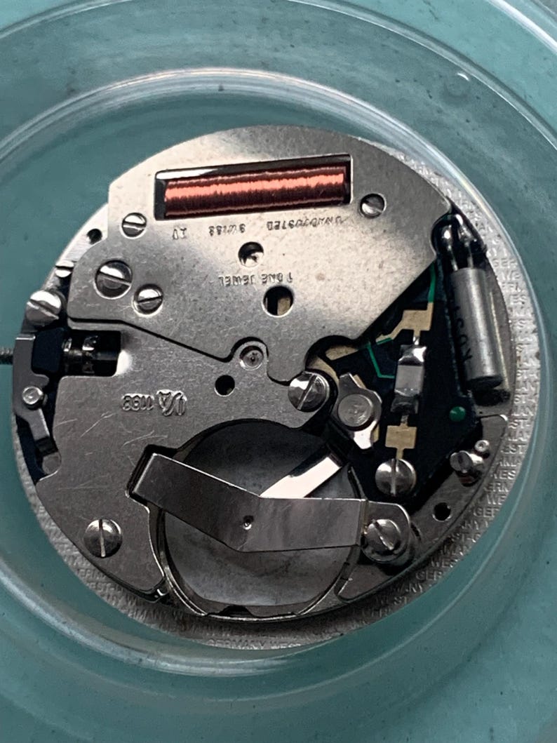 Op de afbeelding: Close-up van een uurwerk met zilverkleurige metalen componenten. Het uurwerk heeft een koperkleurige spoel, een groene printplaat en de tekst "SWISS" en "1 ONE JEWEL". Het uurwerk zit in een doorzichtige, ronde container.