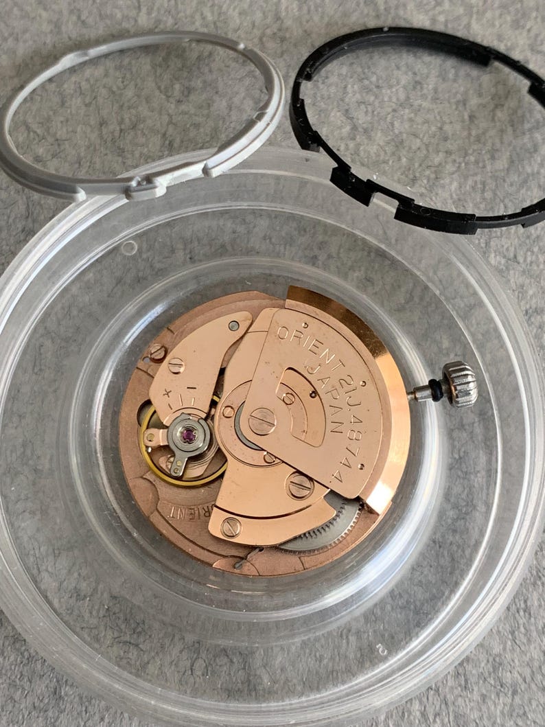 Puede incluir: Un movimiento de reloj desmontado con un mecanismo de color cobre, un recipiente de pl&aacute;stico transparente y dos biseles negros y plateados. El mecanismo del reloj tiene el texto "ORIENT JAPAN 21 JEWELS 46744A".
