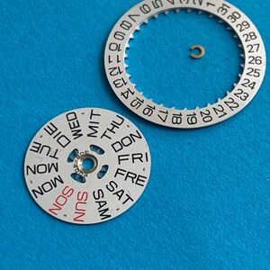 May include: Watch parts on a blue background. The image shows a silver-colored date wheel with numbers 1-31 and a day wheel with the days of the week. A small gold-colored ring is also present. The day wheel has "SUN" in red.