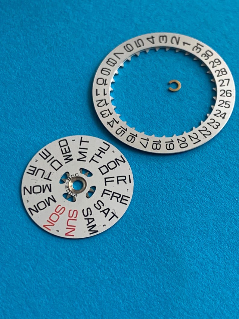 May include: Watch parts on a blue background. The image shows a silver-colored date wheel with numbers 1-31 and a day wheel with the days of the week. A small gold-colored ring is also present. The day wheel has "SUN" in red.