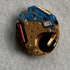 May include: A detailed view of a watch movement featuring gold-toned parts and a blue circuit board. The text "UNADJUSTED SWISS V8" and "ELEVEN JEWELS" are visible. The mechanism includes a small red coil and a silver battery.