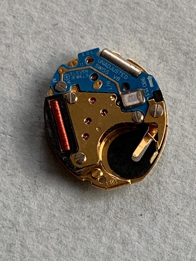 May include: A detailed view of a watch movement featuring gold-toned parts and a blue circuit board. The text "UNADJUSTED SWISS V8" and "ELEVEN JEWELS" are visible. The mechanism includes a small red coil and a silver battery.