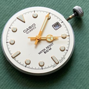 K&ouml;nnte beinhalten: Ein Casio Quartz Zifferblatt mit wei&szlig;em Zifferblatt, goldenen Zeigern und einem Datumsfenster mit der Zahl 26. Das Zifferblatt hat goldene Stundenmarkierungen und den Text "WATER RESIST 50M". Die Krone der Uhr ist sichtbar.