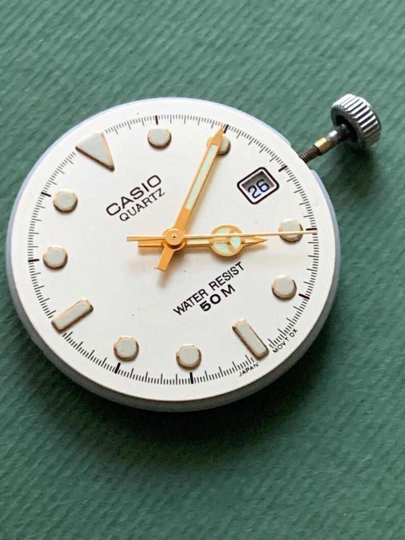 K&ouml;nnte beinhalten: Ein Casio Quartz Zifferblatt mit wei&szlig;em Zifferblatt, goldenen Zeigern und einem Datumsfenster mit der Zahl 26. Das Zifferblatt hat goldene Stundenmarkierungen und den Text "WATER RESIST 50M". Die Krone der Uhr ist sichtbar.