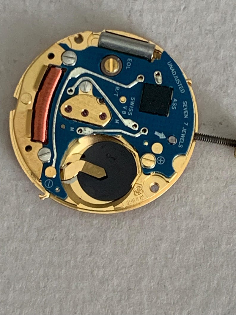 May include: A close-up of a watch movement with a gold-colored frame and a blue circuit board. The board has silver and gold components, including a battery, and the text "UNADJUSTED" and "SEVEN 7 JEWELS".