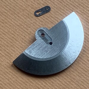 Pu&ograve; includere: Primo piano di due componenti metallici di orologio. Uno &egrave; un pezzo semicircolare con un foro e un bordo nero. L'altro &egrave; un piccolo pezzo oblungo scuro con due fori. Entrambi i pezzi sono su una superficie marrone chiaro.