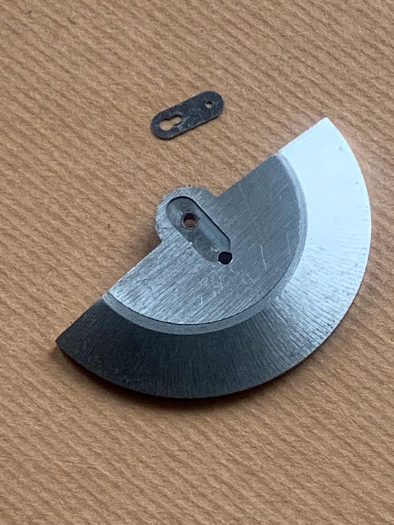 Pu&ograve; includere: Primo piano di due componenti metallici di orologio. Uno &egrave; un pezzo semicircolare con un foro e un bordo nero. L'altro &egrave; un piccolo pezzo oblungo scuro con due fori. Entrambi i pezzi sono su una superficie marrone chiaro.