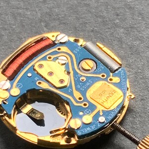 May include: Close-up of a watch movement with a blue circuit board and gold-colored components. The watch mechanism has the text "ZENITH" and "SWISS" printed on it. The watch has a screw and a small, clear window.