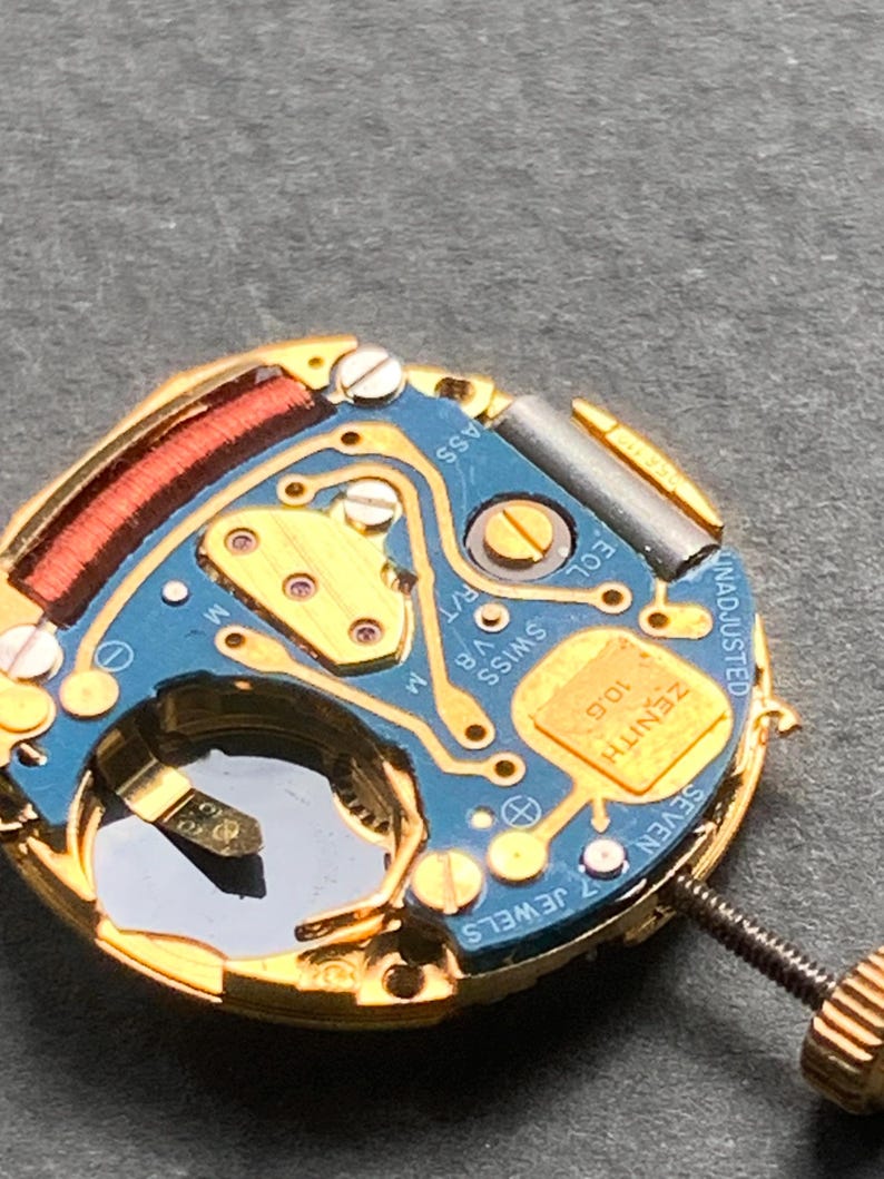 May include: Close-up of a watch movement with a blue circuit board and gold-colored components. The watch mechanism has the text "ZENITH" and "SWISS" printed on it. The watch has a screw and a small, clear window.