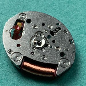 May include: Close-up of a watch movement with a silver-colored metal base. The movement features small gears, screws, and a copper-colored coil. The background is a solid teal color.