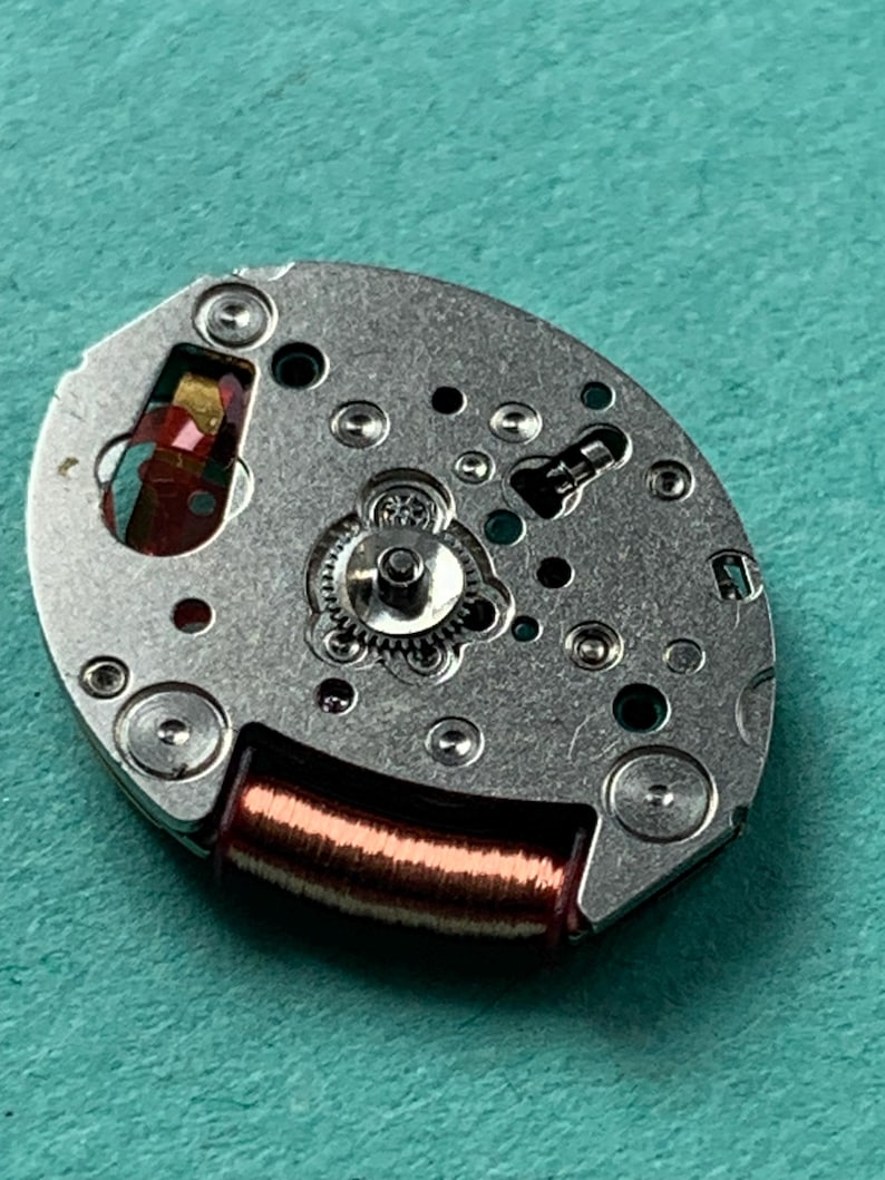 May include: Close-up of a watch movement with a silver-colored metal base. The movement features small gears, screws, and a copper-colored coil. The background is a solid teal color.