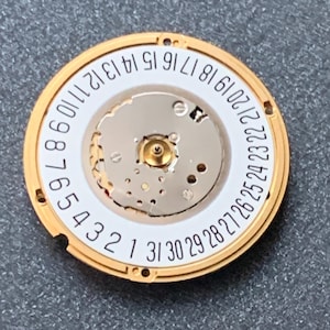 May include: A gold-toned watch movement component with a white inner dial and a ring of numbers from 1 to 31. The component has intricate mechanical details and is designed for a watch. The background is a gray surface.