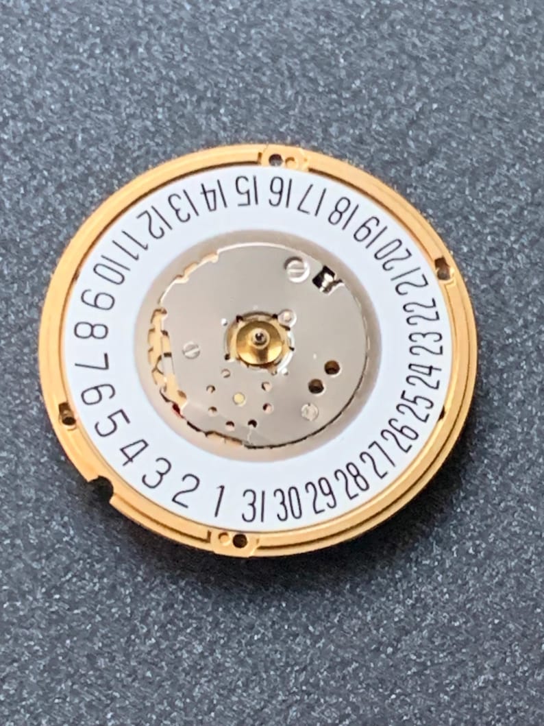 May include: A gold-toned watch movement component with a white inner dial and a ring of numbers from 1 to 31. The component has intricate mechanical details and is designed for a watch. The background is a gray surface.