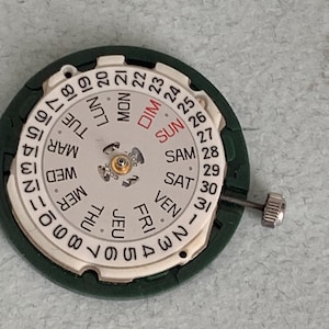 May include: A watch movement with a white dial and a green outer ring. The dial displays the days of the week in English and numbers 1-31. A small screw is attached to the side.