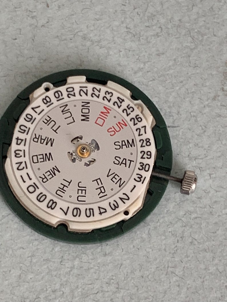 May include: A watch movement with a white dial and a green outer ring. The dial displays the days of the week in English and numbers 1-31. A small screw is attached to the side.