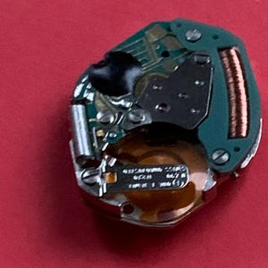 May include: A watch movement with a green circuit board, copper coil, and silver components. The watch mechanism is set against a red background. The watch movement has small screws and a black component.