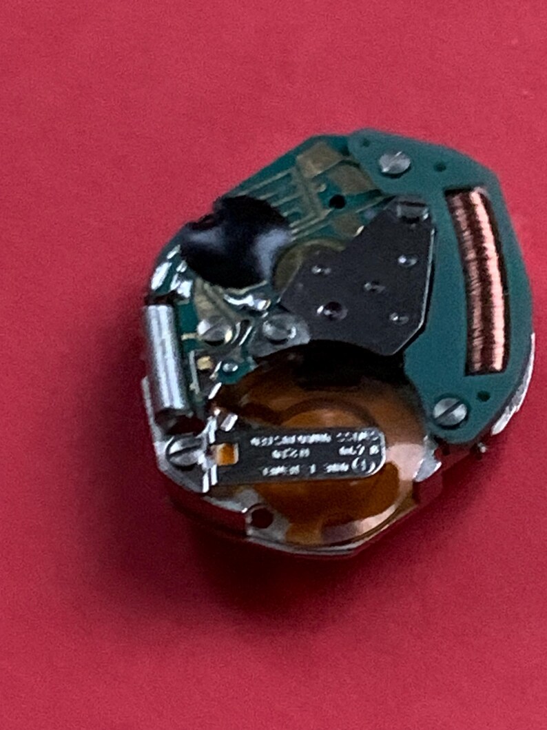 May include: A watch movement with a green circuit board, copper coil, and silver components. The watch mechanism is set against a red background. The watch movement has small screws and a black component.