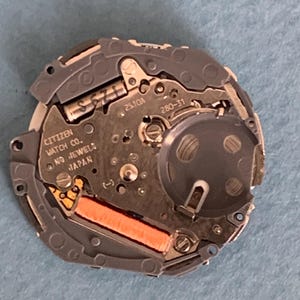 May include: A close-up of a Citizen watch movement, featuring intricate gears, circuits, and a battery compartment. The watch mechanism is primarily silver and gray, with orange and gold accents. The text "CITIZEN WATCH CO. NO JEWELS JAPAN" is visible.