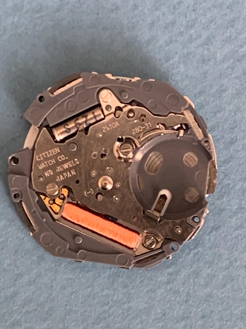 May include: A close-up of a Citizen watch movement, featuring intricate gears, circuits, and a battery compartment. The watch mechanism is primarily silver and gray, with orange and gold accents. The text "CITIZEN WATCH CO. NO JEWELS JAPAN" is visible.