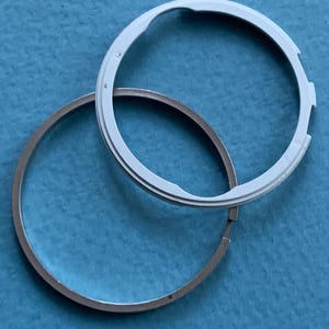 May include: Two circular watch components. One is a silver-coloured ring, and the other is a white ring with a notched design. The components are positioned on a blue surface, with the white ring overlapping the silver ring.