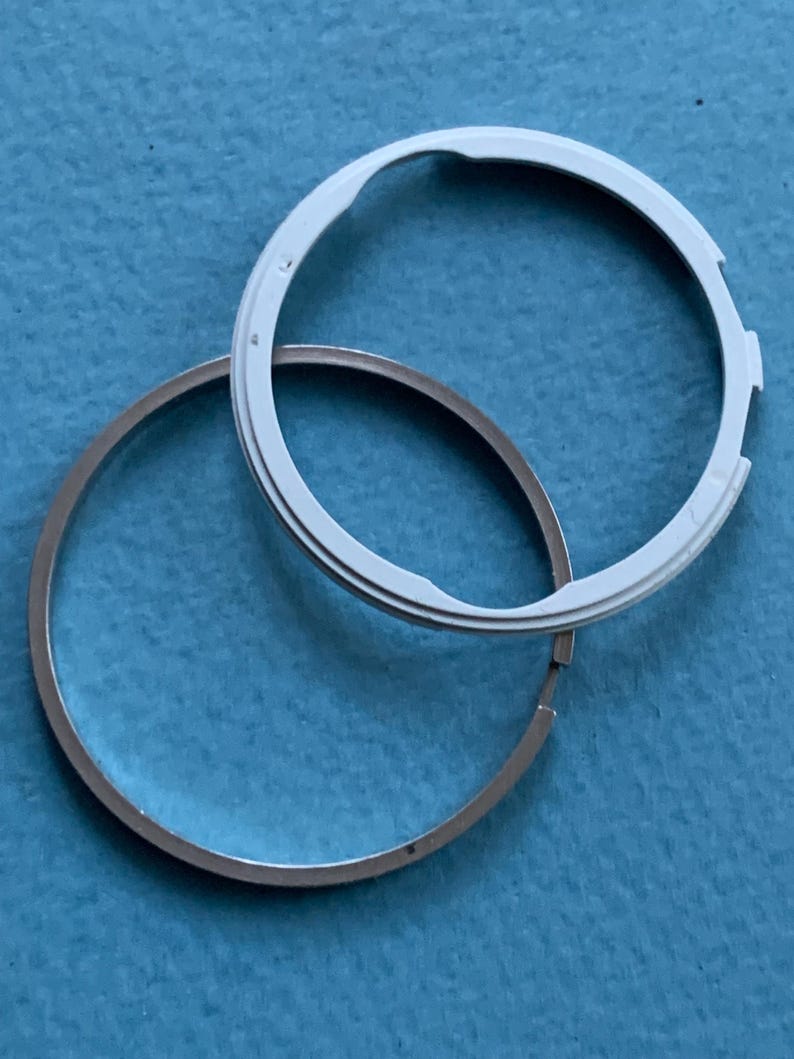 May include: Two circular watch components. One is a silver-coloured ring, and the other is a white ring with a notched design. The components are positioned on a blue surface, with the white ring overlapping the silver ring.