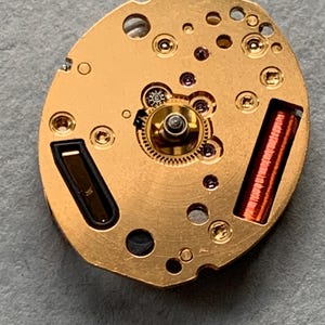 May include: Close-up of a watch movement with a gold-coloured base. The movement features various gears, screws, and a copper-coloured coil. The intricate design showcases the internal mechanics of a timepiece.