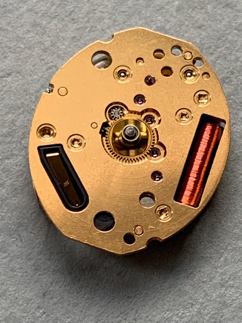 May include: Close-up of a watch movement with a gold-coloured base. The movement features various gears, screws, and a copper-coloured coil. The intricate design showcases the internal mechanics of a timepiece.