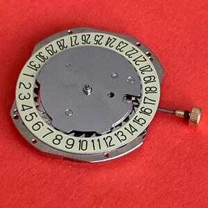 K&ouml;nnte beinhalten: Ein Uhrwerk mit silberfarbenem Zifferblatt und einem wei&szlig;en Ring mit den Tagen des Monats, nummeriert von 1 bis 31. Eine kleine goldfarbene Krone und eine Schraube sind sichtbar. Der Hintergrund ist rot.