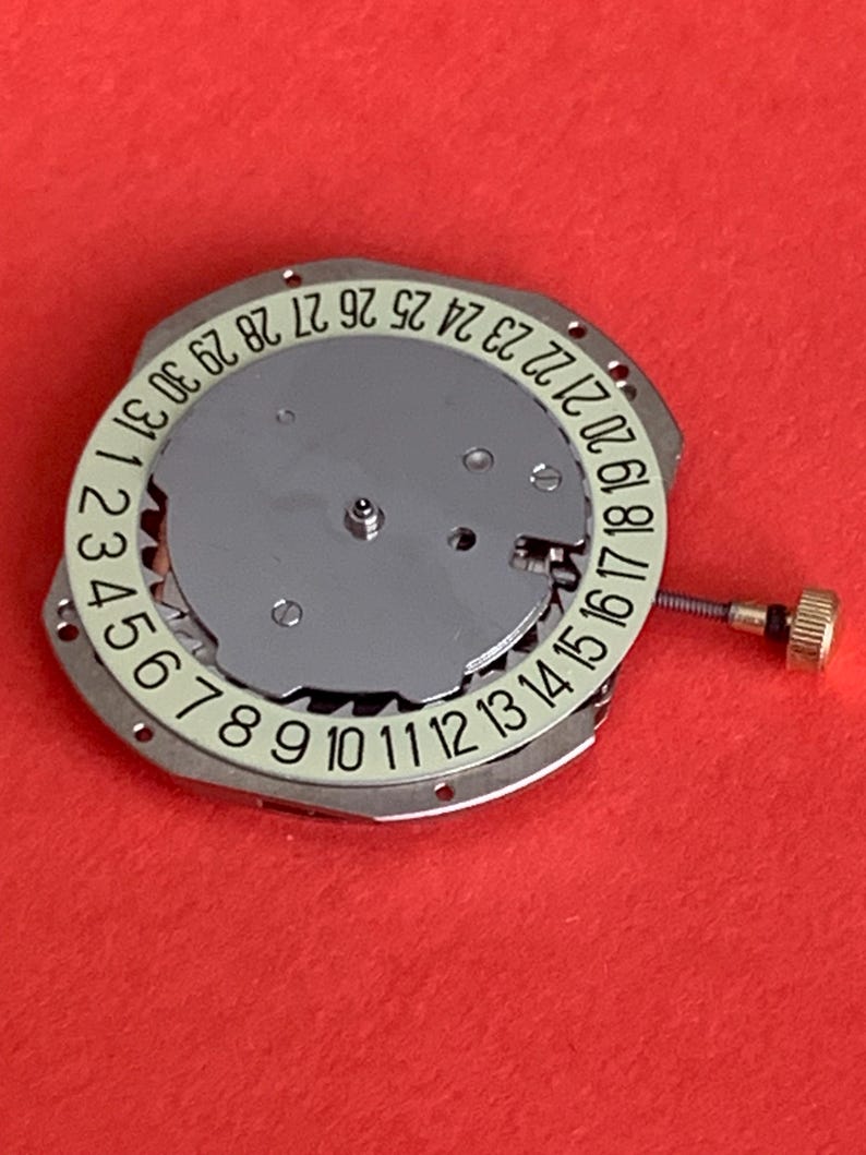 K&ouml;nnte beinhalten: Ein Uhrwerk mit silberfarbenem Zifferblatt und einem wei&szlig;en Ring mit den Tagen des Monats, nummeriert von 1 bis 31. Eine kleine goldfarbene Krone und eine Schraube sind sichtbar. Der Hintergrund ist rot.