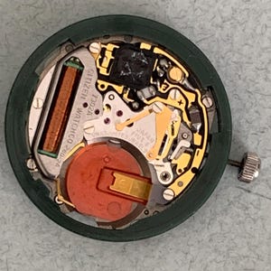 May include: A Citizen watch movement, model 7302A, with a green outer ring. The internal components include a copper coil, a red battery, and gold-colored gears. The watch movement is labeled "Japan Five Jewels".