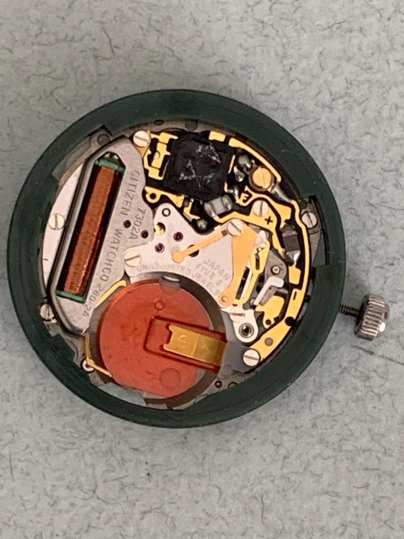 May include: A Citizen watch movement, model 7302A, with a green outer ring. The internal components include a copper coil, a red battery, and gold-colored gears. The watch movement is labeled "Japan Five Jewels".