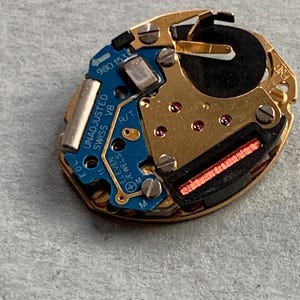 May include: A close-up of a watch movement with gold-coloured metal components and a blue circuit board. The board has the text "UNADJUSTED SWISS V8 ELEVEN JEWELS". A black battery and red components are also visible.