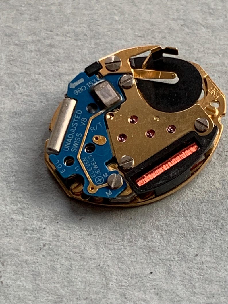May include: A close-up of a watch movement with gold-coloured metal components and a blue circuit board. The board has the text "UNADJUSTED SWISS V8 ELEVEN JEWELS". A black battery and red components are also visible.
