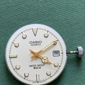 K&ouml;nnte beinhalten: Ein Casio Quartz Zifferblatt mit cremefarbenem Zifferblatt, goldenen Zeigern und Markierungen. Das Zifferblatt zeigt das Datum 26 und den Text "WATER RESIST 50 M". Das Zifferblatt liegt auf einer gr&uuml;nen Oberfl&auml;che.