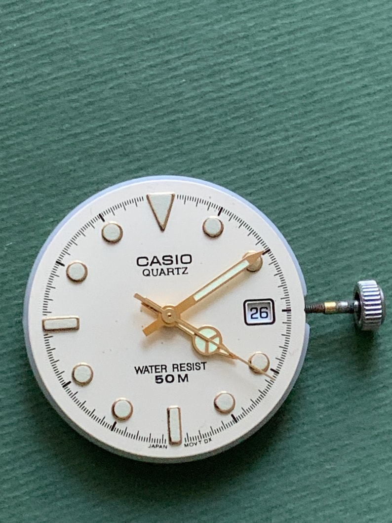 K&ouml;nnte beinhalten: Ein Casio Quartz Zifferblatt mit cremefarbenem Zifferblatt, goldenen Zeigern und Markierungen. Das Zifferblatt zeigt das Datum 26 und den Text "WATER RESIST 50 M". Das Zifferblatt liegt auf einer gr&uuml;nen Oberfl&auml;che.