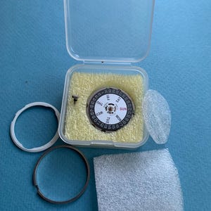 May include: A watch movement with a black and white dial, showing days of the week and numbers. It is in a clear plastic case with yellow foam padding. Additional watch parts, including a metal ring and a clear plastic ring, are also present.