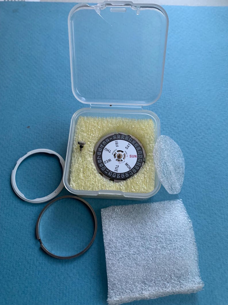 May include: A watch movement with a black and white dial, showing days of the week and numbers. It is in a clear plastic case with yellow foam padding. Additional watch parts, including a metal ring and a clear plastic ring, are also present.