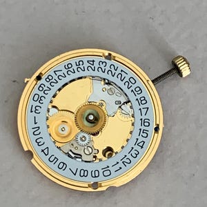May include: Close-up of a watch movement with gold-colored gears and a light blue date dial. The dial displays numbers from 1 to 31. A small screw and crown are attached to the side.