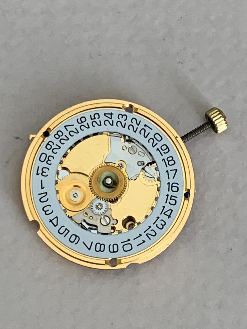 May include: Close-up of a watch movement with gold-colored gears and a light blue date dial. The dial displays numbers from 1 to 31. A small screw and crown are attached to the side.