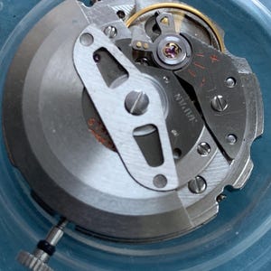 May include: Detailed view of a watch movement, showcasing its complex internal workings. The mechanism comprises silver-toned metal components, a gold-coloured ring, and the word "JAPAN" inscribed on a part. A small, textured knob is present.