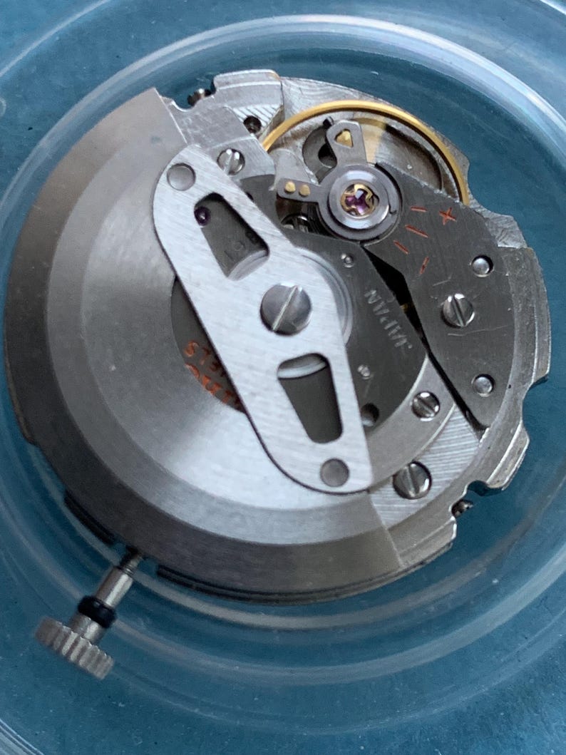 May include: Detailed view of a watch movement, showcasing its complex internal workings. The mechanism comprises silver-toned metal components, a gold-coloured ring, and the word "JAPAN" inscribed on a part. A small, textured knob is present.