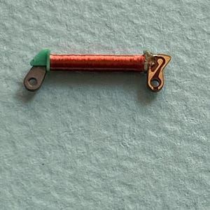 May include: Close-up of a small, copper-colored coil with green and gold-colored end pieces. The coil is tightly wound and has a small hole on each end. The object is likely an electronic component.