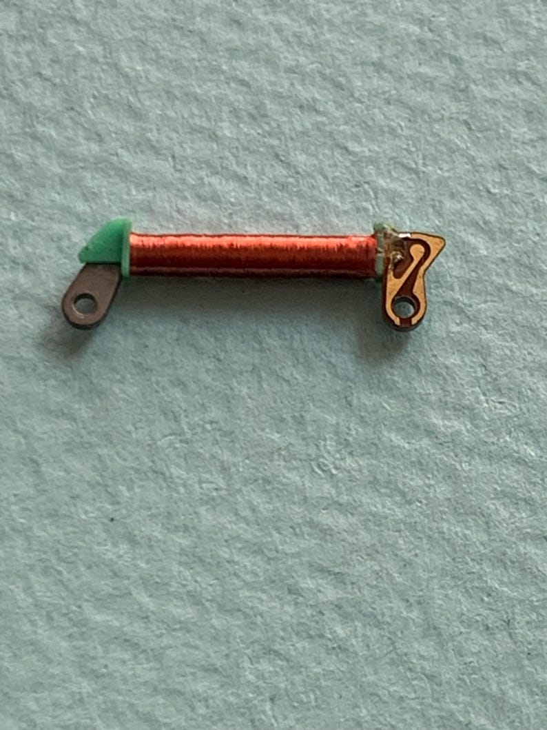 May include: Close-up of a small, copper-colored coil with green and gold-colored end pieces. The coil is tightly wound and has a small hole on each end. The object is likely an electronic component.